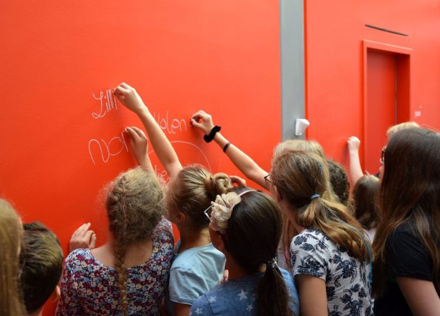 Ein Gruppe von Mädchen steht vor einer roten Wand. Einige Mädchen schreiben mit Kreide an die Wand.